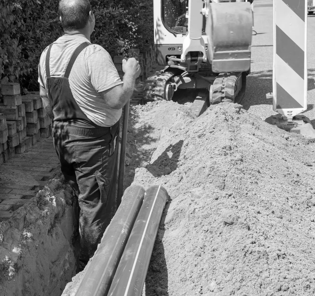 Arbeiter an einem offenen Leitungsgraben mit verlegten PE-Rohren, im Hintergrund ein Minibagger